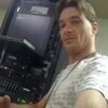 A person wearing a light-colored shirt and lanyard takes a selfie in front of server racks and electronic equipment, possibly working with a Hosted VoIP Switch in a technical or server room environment.