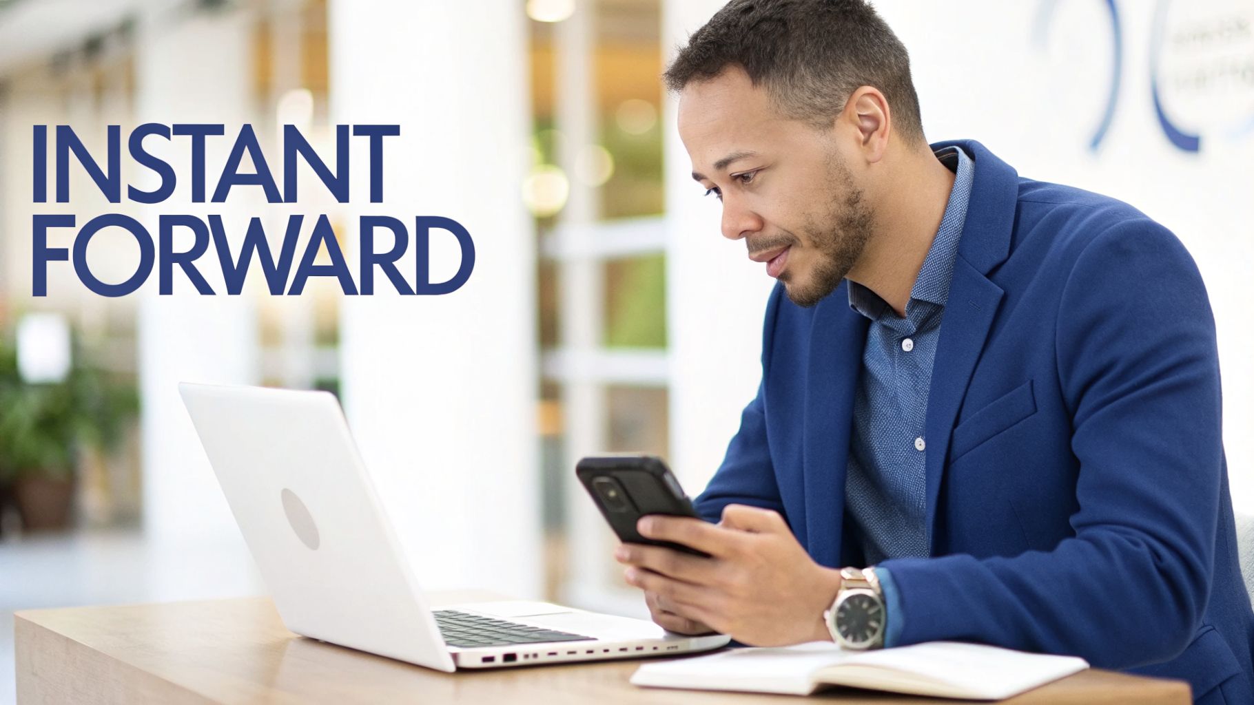 A man in a blue suit looks at his smartphone while sitting at a desk with a laptop.