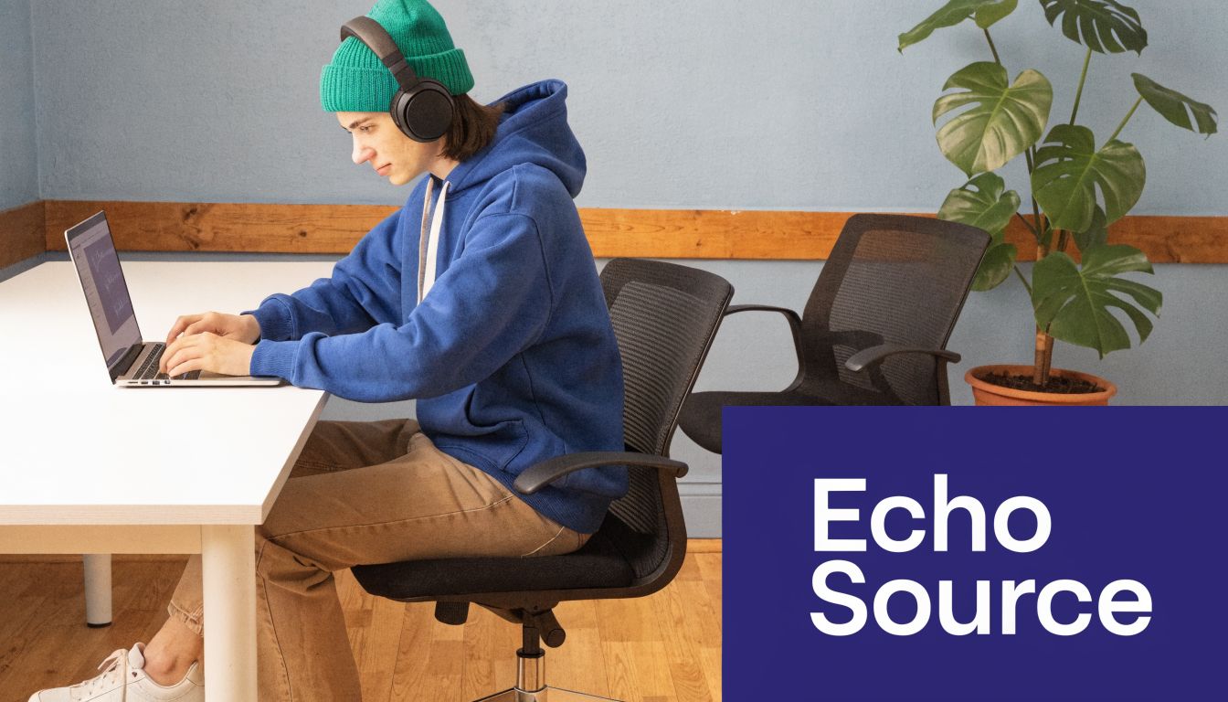 A young person wearing a green beanie and blue hoodie working on a laptop at a desk.