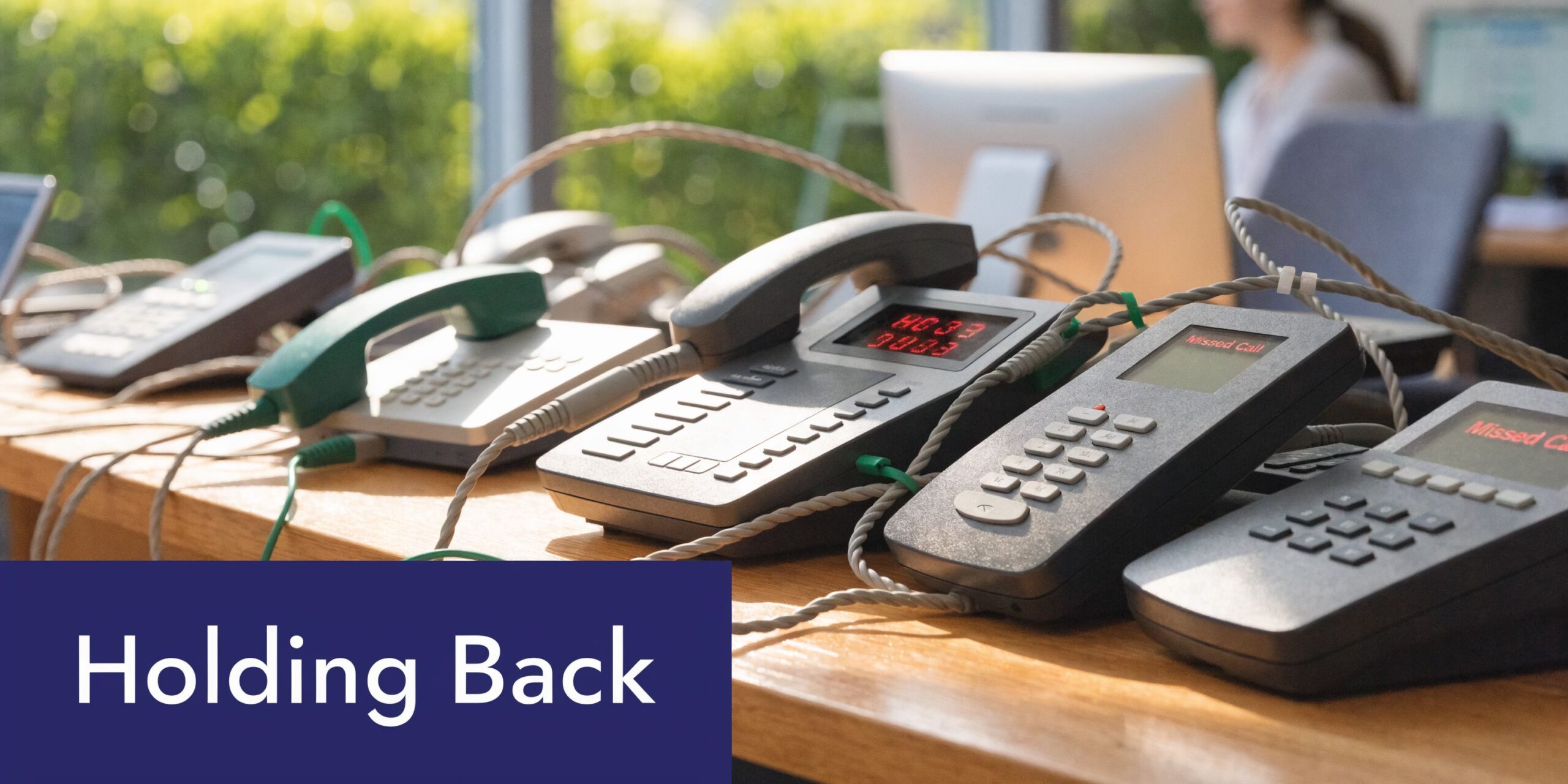A row of office VOIP phones lined up on a wooden desk in a busy workplace.