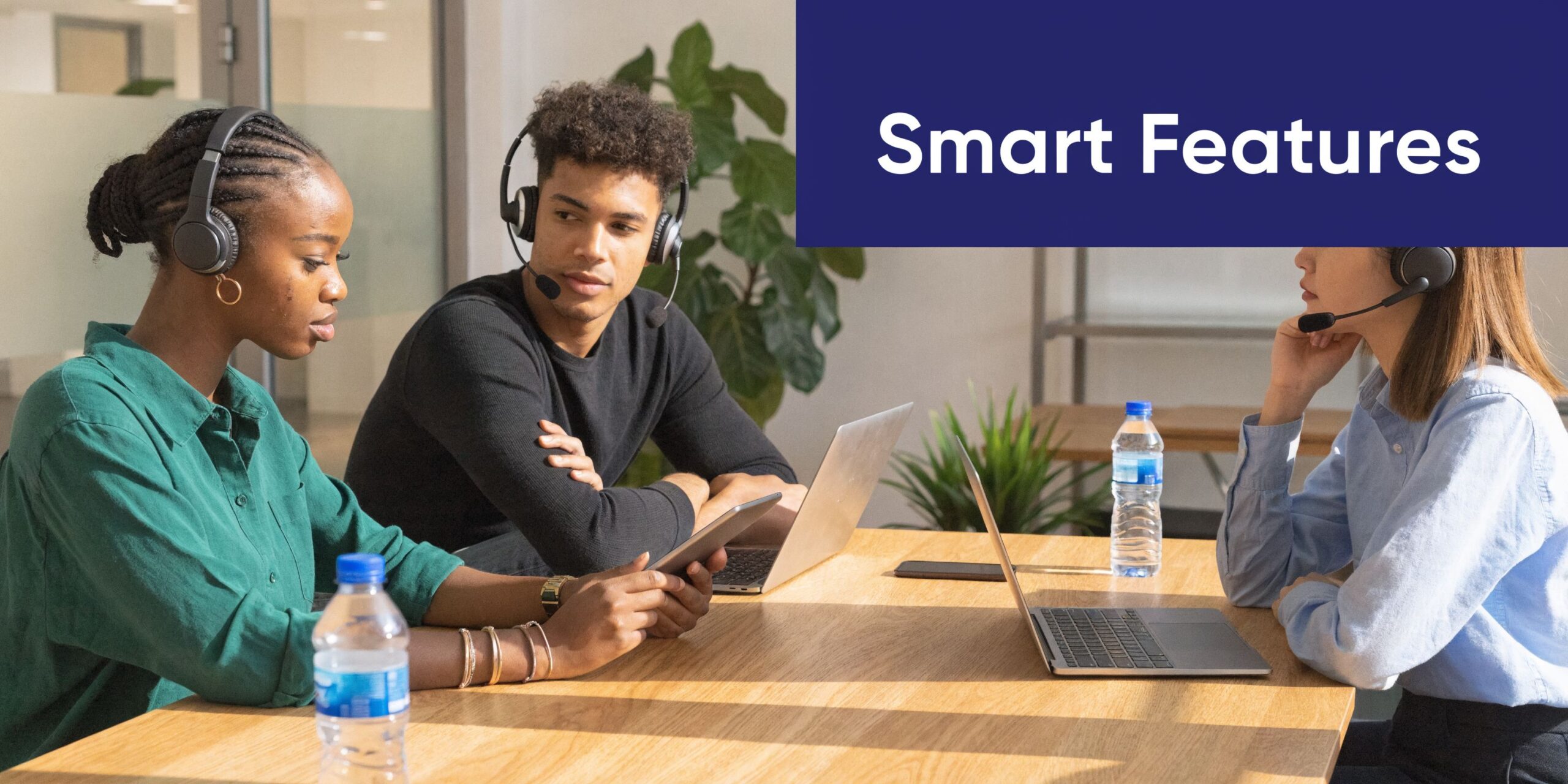 Three professionals wearing headsets collaborating in a modern office space while using laptops and a tablet device.