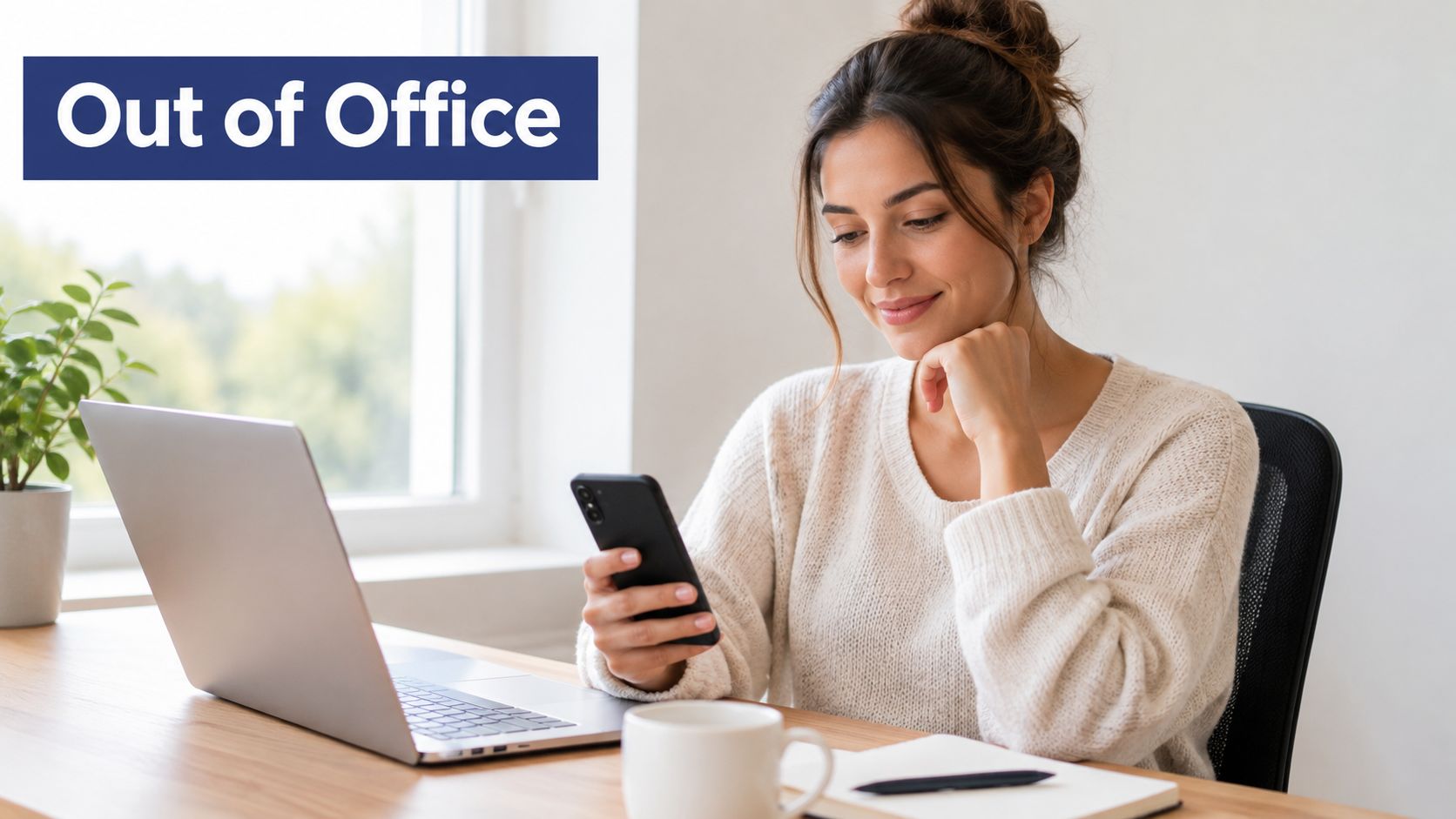 A woman sits at a wooden desk with a laptop, looking at her smartphone while smiling.